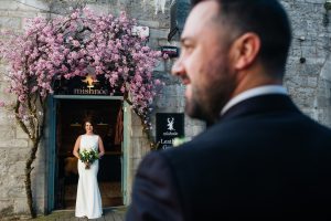 Bride under pink blossom with Groom looking on